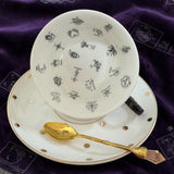 Teacup with decorative patterns on a matching saucer and a gold spoon on a purple fabric background.