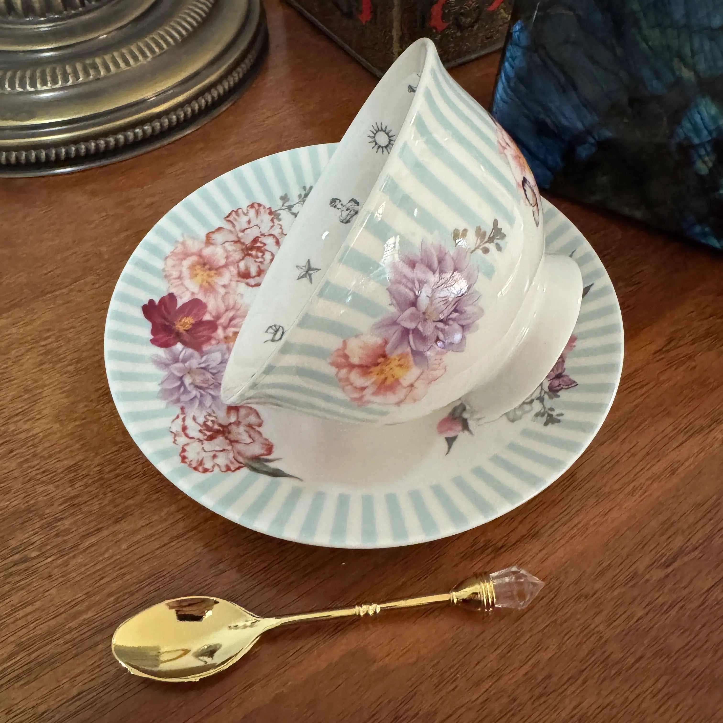 A white porcelain teacup and saucer set with light blue stripes and pink floral patterns, shown on a wooden table with a gold spoon.