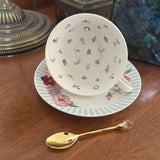 A white ceramic teacup and saucer set on a wooden table, decorated with small black doodle-like motifs inside the cup and a floral border with pale blue stripes around the rim. A small gold spoon with a clear crystal handle rests in front.