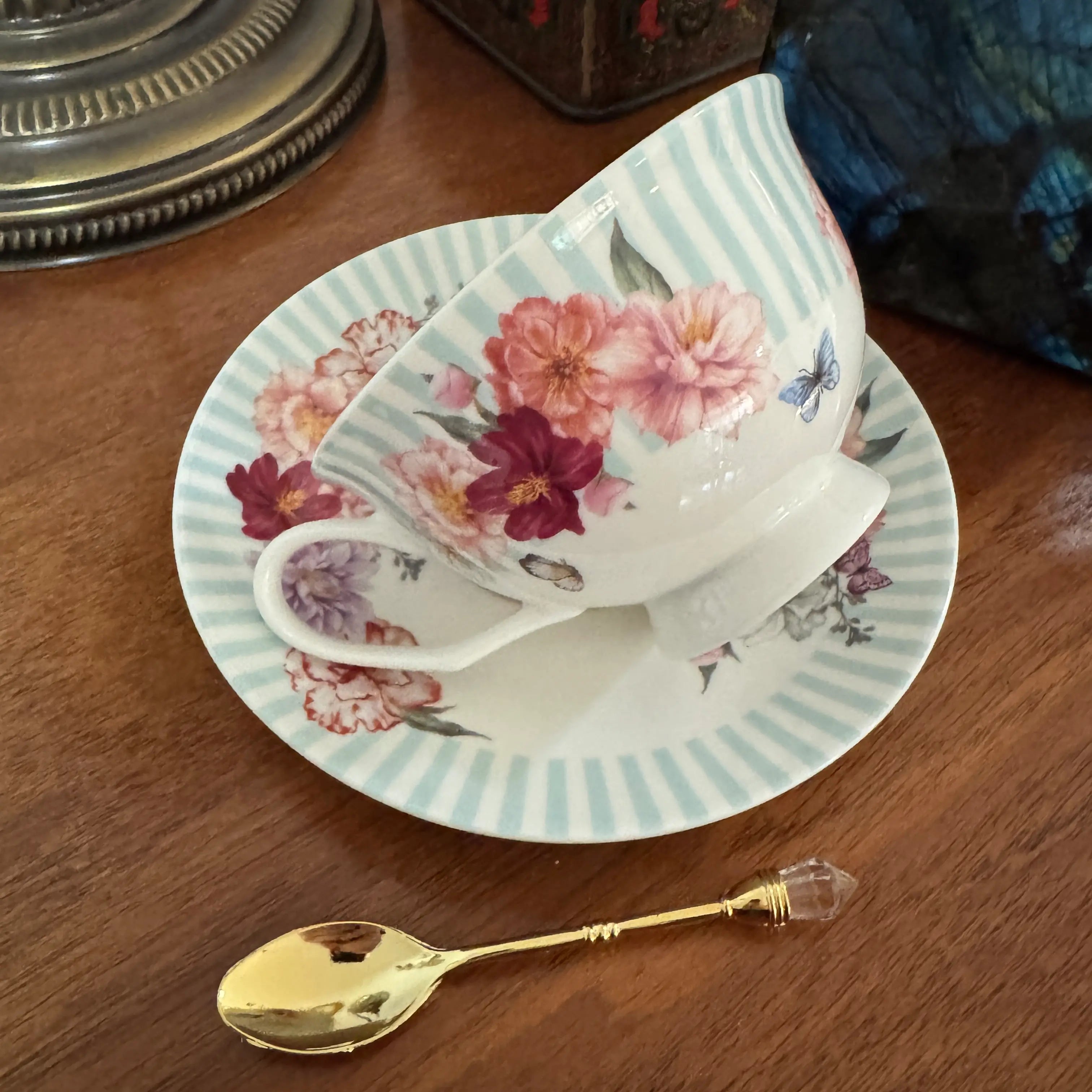 A white porcelain teacup and saucer set with a floral print in pink, red, and purple, featuring light blue-green stripes on the saucer rim. A small gold spoon with a crystal handle rests in front on a wooden table.