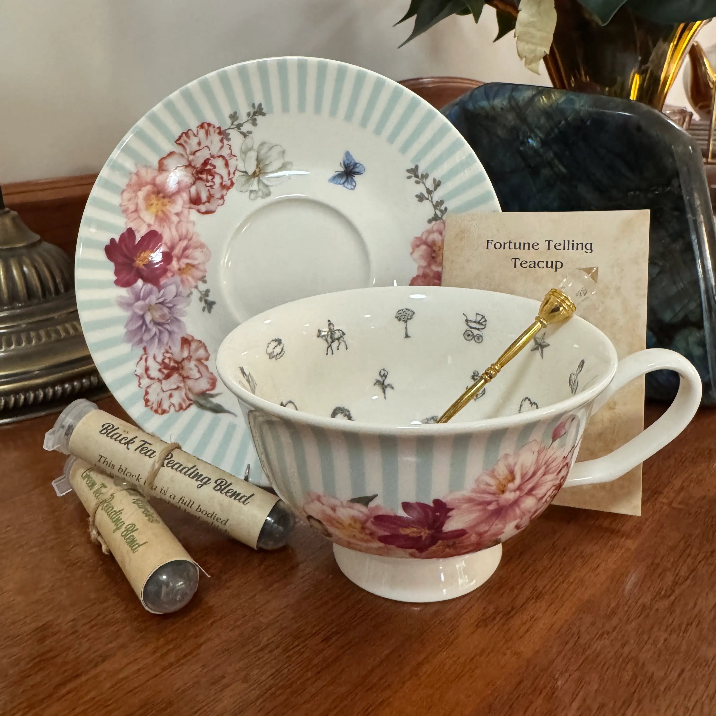 A ceramic teacup and saucer set displayed on a wooden table, featuring pale blue stripes, pink and red floral motifs, and small black-and-white whimsical illustrations. A gold spoon rests inside the cup, with a small card reading 'Fortune Telling Teacup' and two wrapped tea heating sticks nearby.