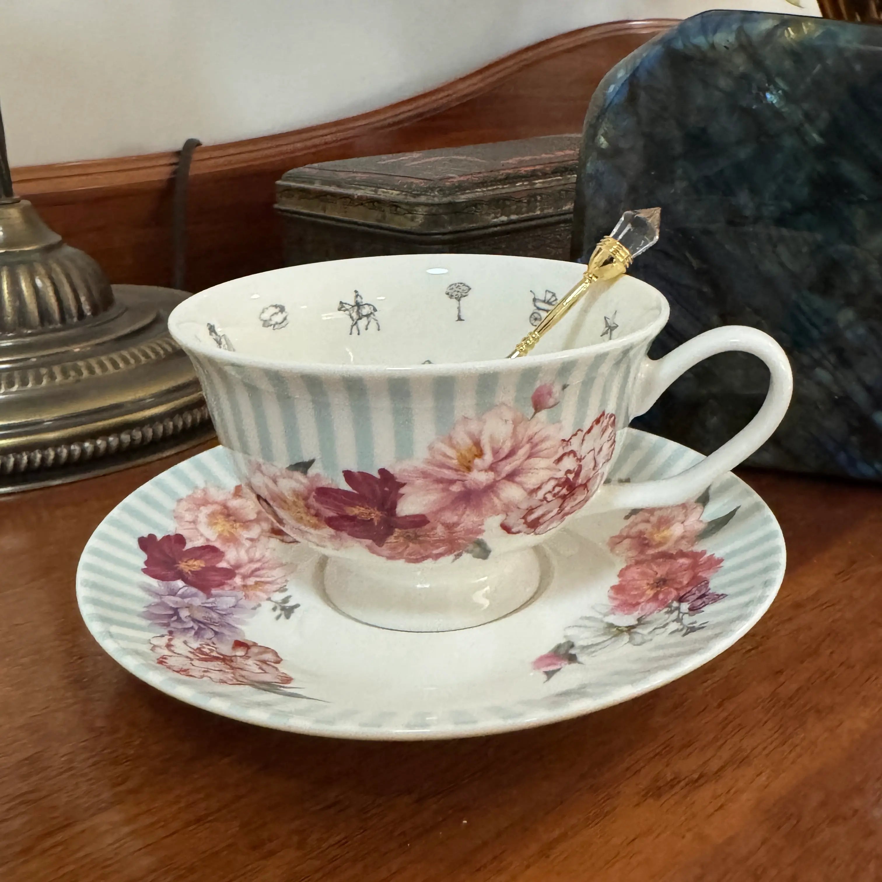 A white porcelain teacup and saucer set with light blue vertical stripes and pink floral patterns, featuring a small gold spoon inside the cup, placed on a wooden table.
