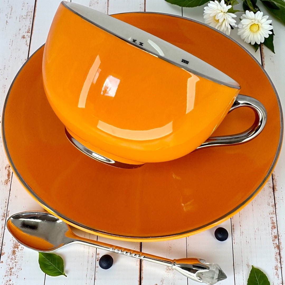 Right side view of the glossy orange teacup sitting on its saucer, highlighting the silver handle and rim details.