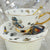 A short video showing a white porcelain teacup and saucer with a steampunk-style cat design, slowly rotating on a display stand. The cup and saucer have gold rims and a matching gold spoon is inside the cup. The background is a white surface decorated with pearl-like beads.