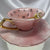 Pink and white marbled teacup and saucer set with a gold handle and rim, featuring internal black symbols for tasseography or tea leaf reading.