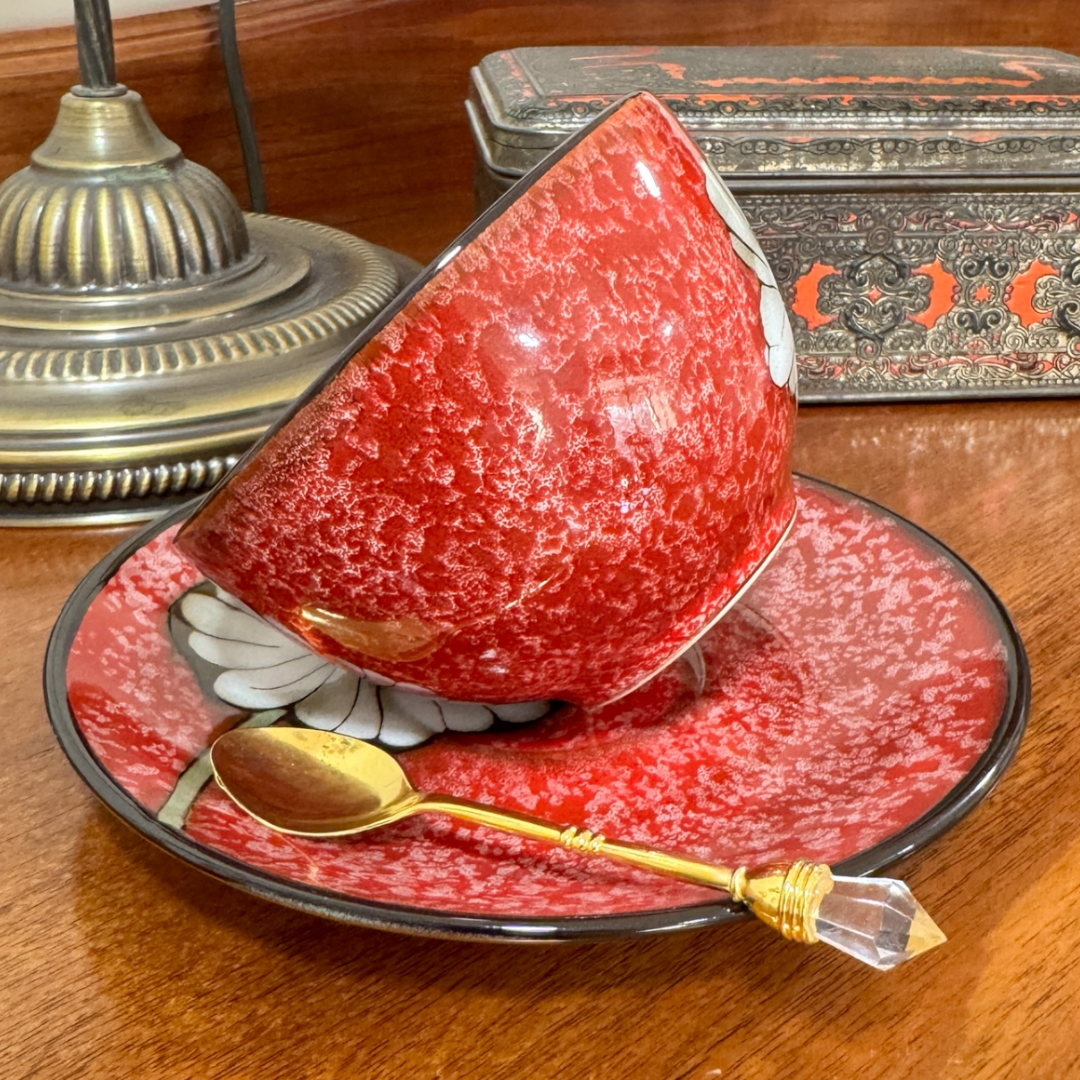 Red ceramic teapot with a matching plate and gold spoon on a wooden surface.