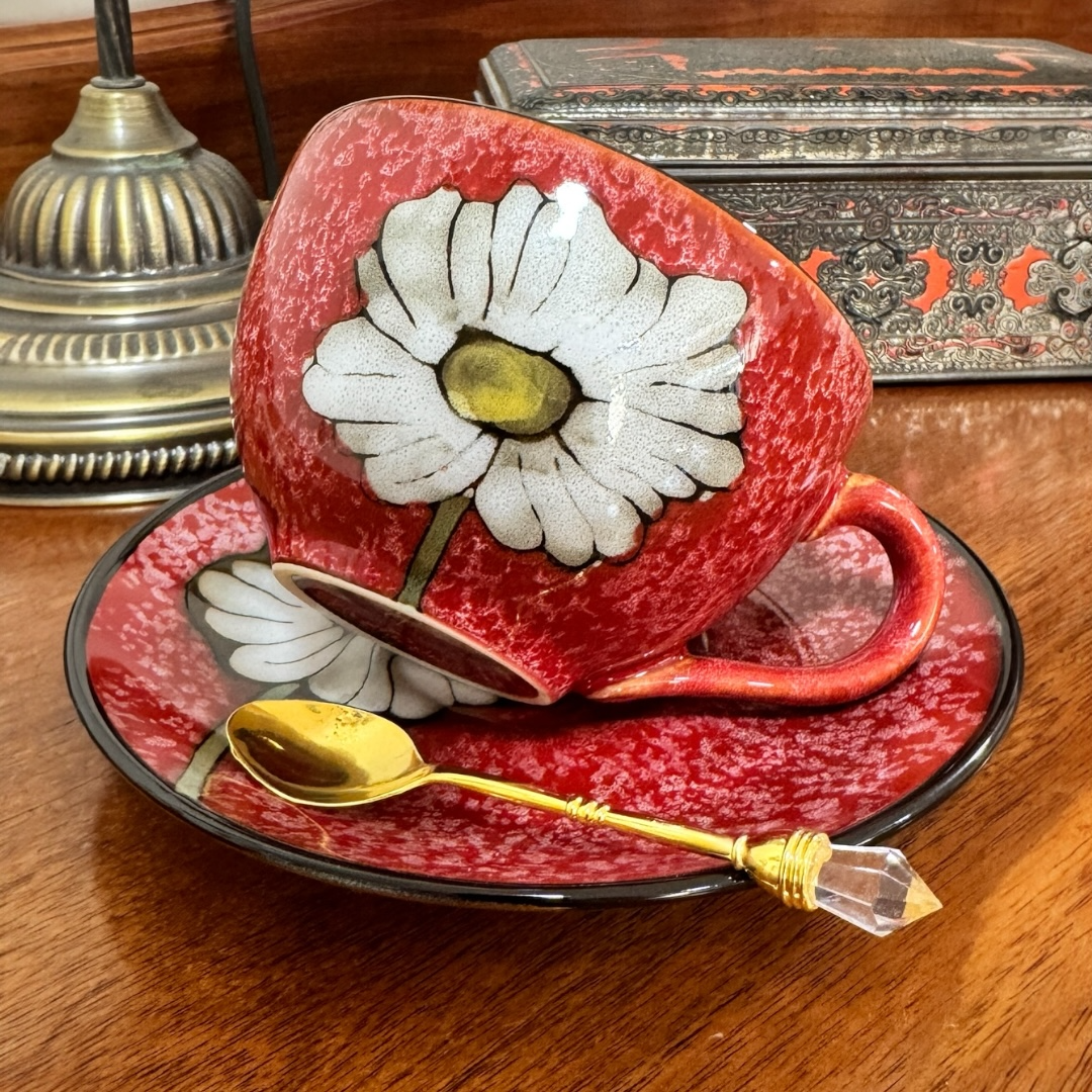 Red teacup with a white flower design on a matching saucer, accompanied by a gold spoon on a wooden surface.