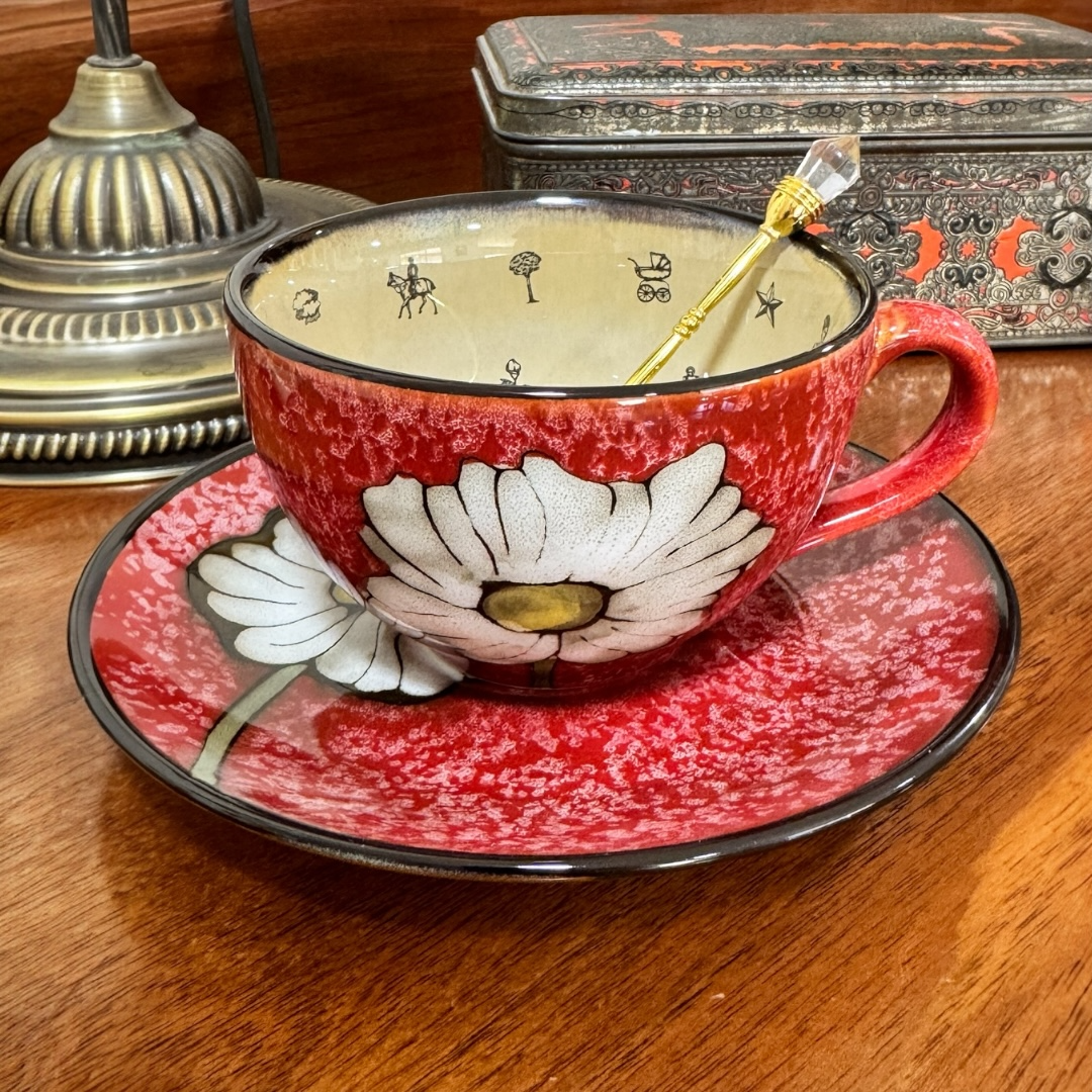 Red floral teacup and saucer with a decorative tin in the background on a wooden surface.