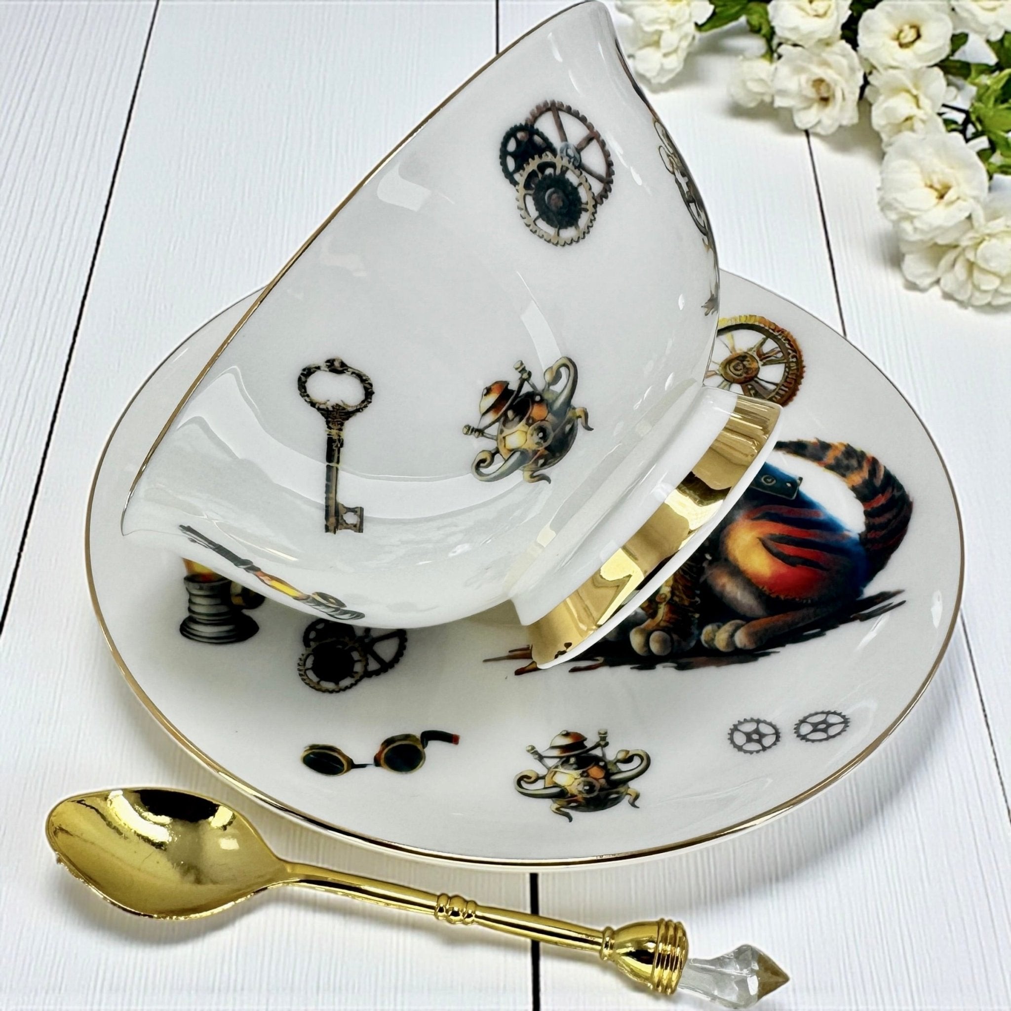 A teacup and saucer set featuring a blue steampunk-style crow in a top hat, with a white porcelain spoon. Two small tubes of tea are also visible.