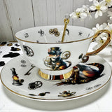A white porcelain teacup with a gold handle and rim, featuring a steampunk-style cat in a top hat and goggles. The saucer shows a similar cat and various steampunk motifs like gears and keys.