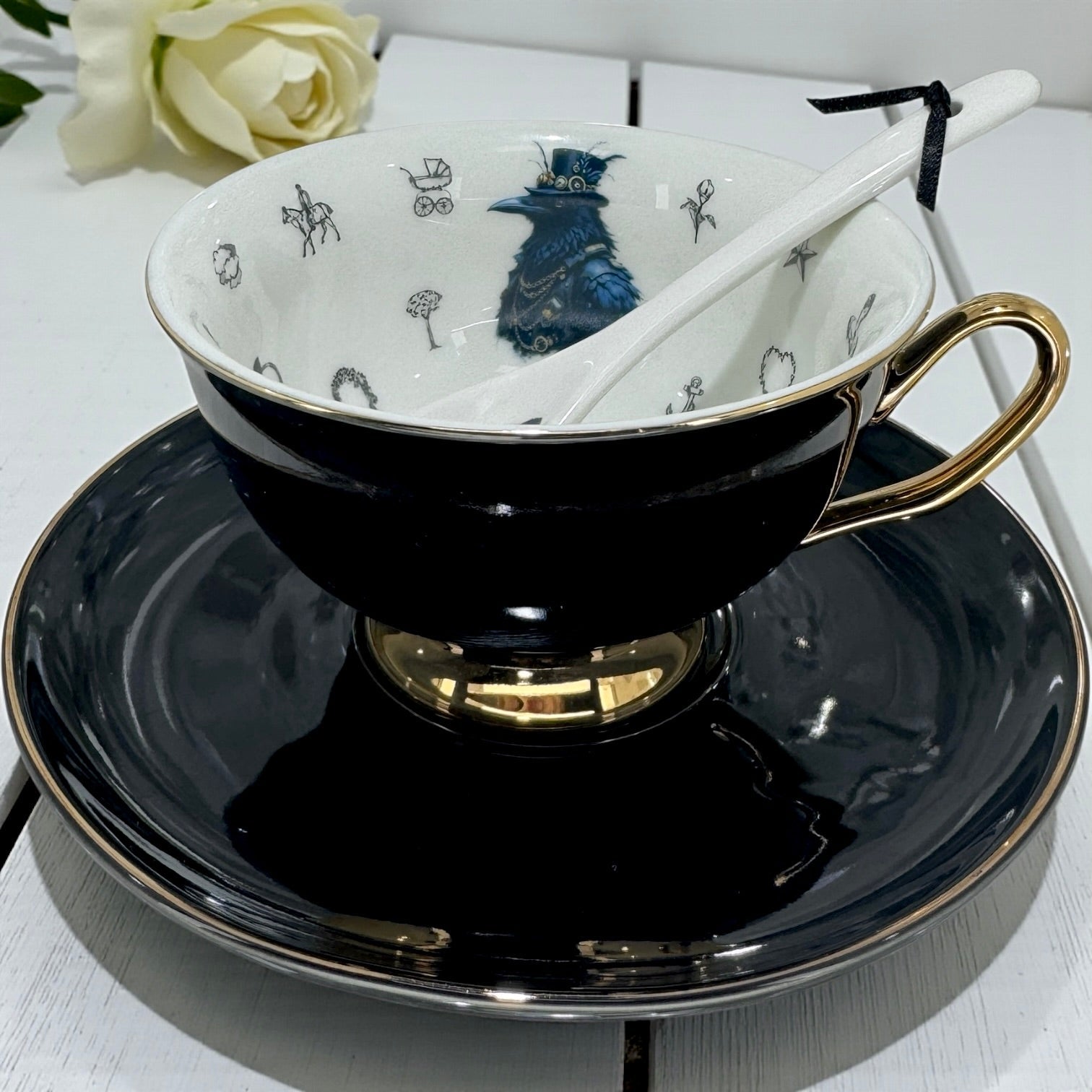 Black teacup with gold handle and saucer on a white surface with a white rose.