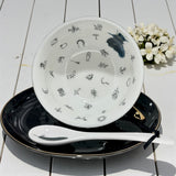 White plate with black patterns on a black plate with a white spoon, on a light wooden surface with flowers.