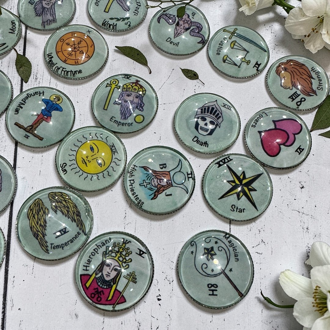 Overhead view of 22 tarot charm casting coins arranged on a rustic surface with white flowers showing individual Major Arcana illustrations under rounded glass tops on metal discs