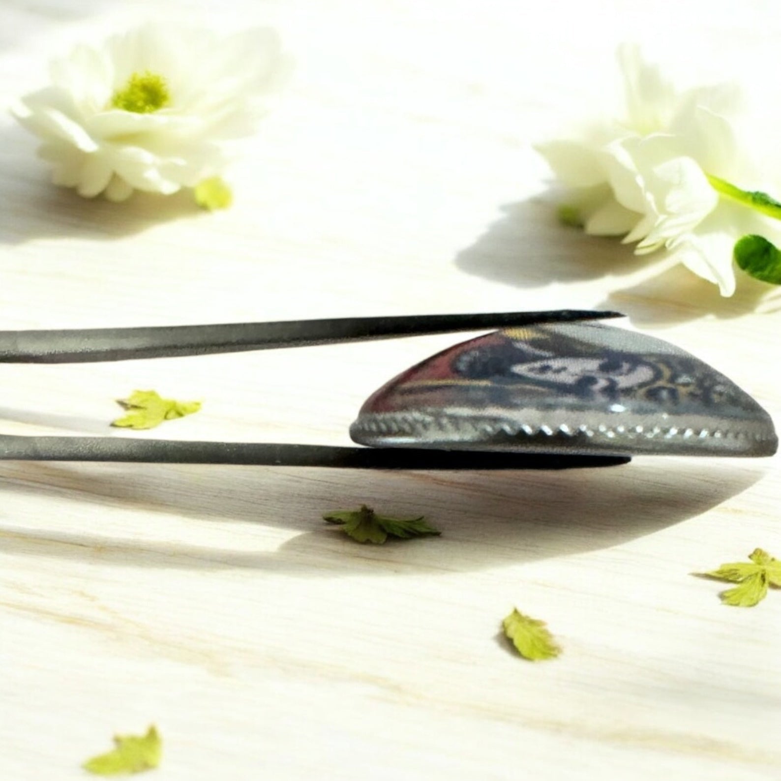 Side profile of a single tarot charm casting coin held in tweezers showing the clear glass dome top and smooth metal backing plate construction