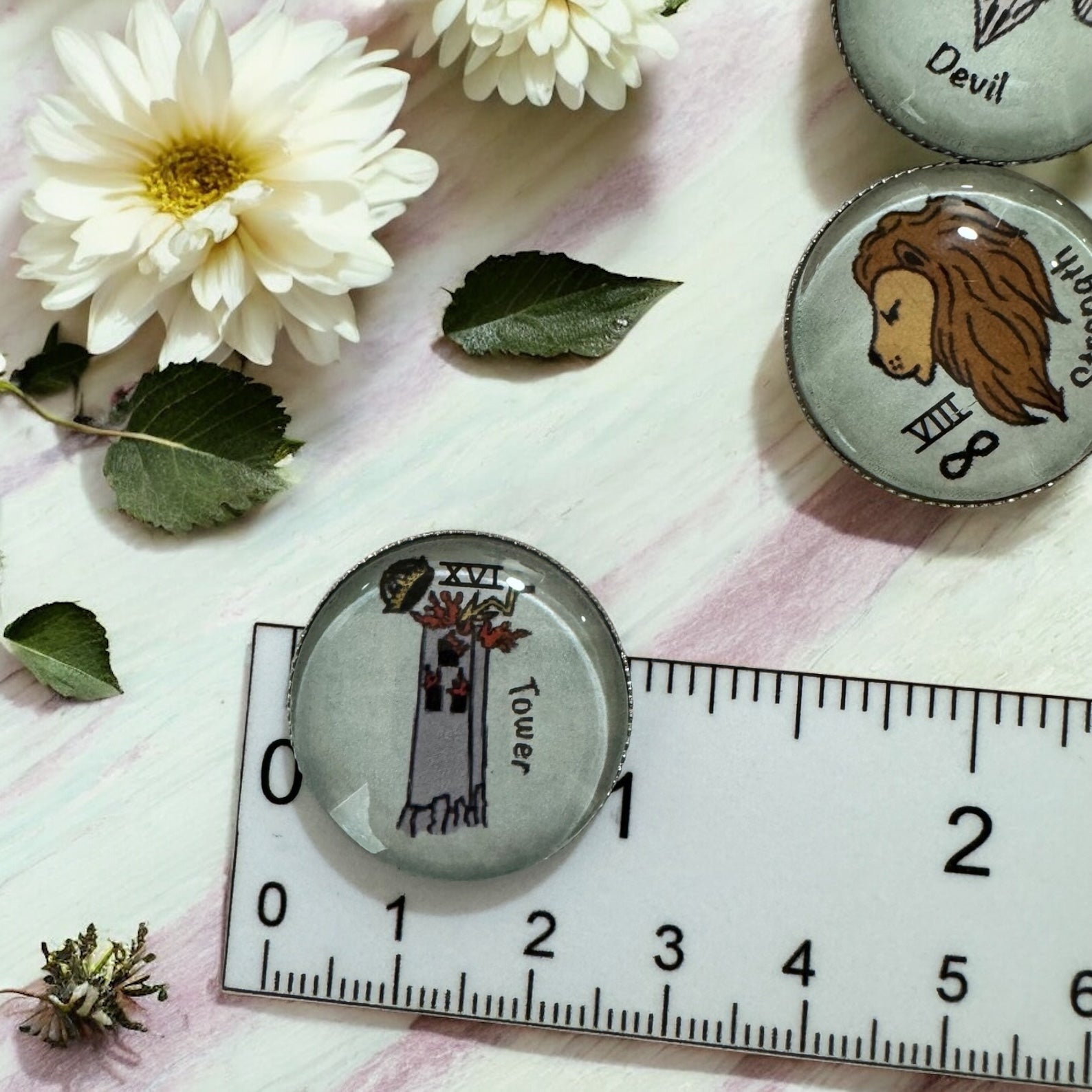 Tarot casting coins shown next to a ruler for scale measuring 25mm diameter each with glass dome top over hand-drawn Tower and Devil card artwork on metal coin base
