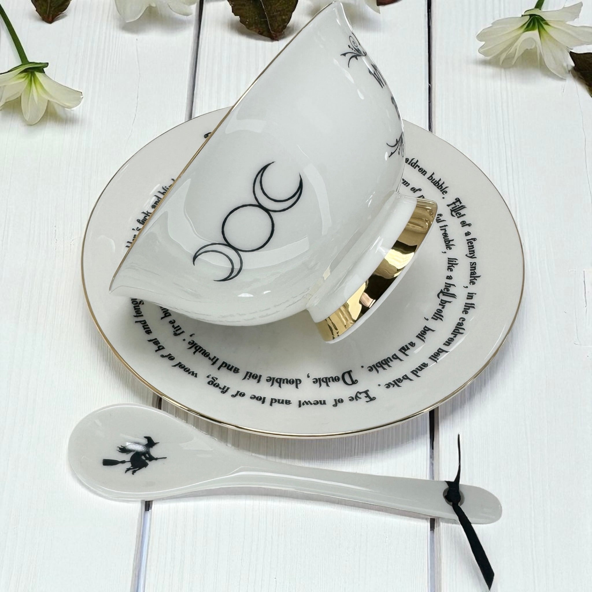 Teacup, saucer, and spoon with black designs on a white wooden surface with flowers.