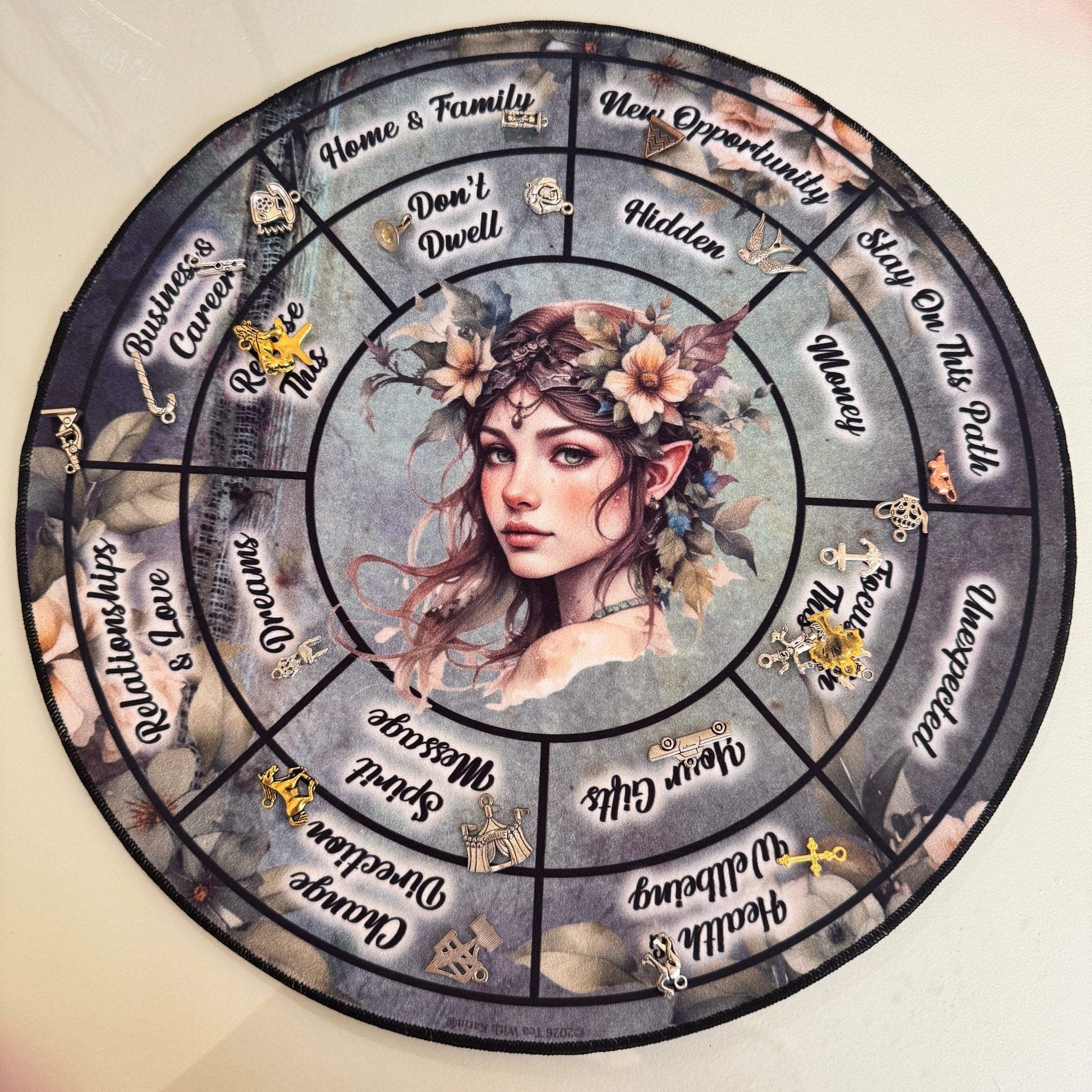 Overhead view of a circular divination mat featuring central elf artwork and silver charms on various life sectors.